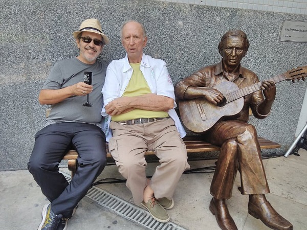 O escritor jorge fernando está assentado junto do compositor Pacífico Mascarenhas, perto da estátua dele, tocando violão